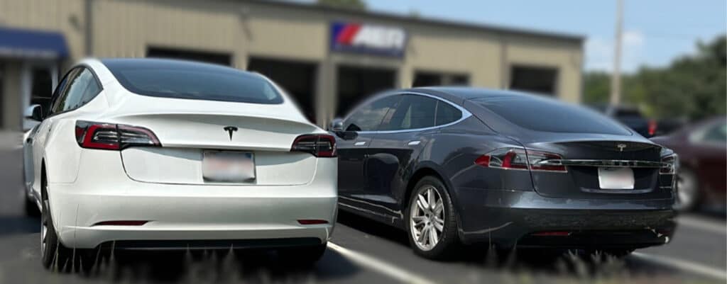 Expert Tesla maintenance services in Franklin, MA with Advanced European Repair. Image of the back of a white Tesla and Gray Tesla parked in the AER shop parking lot after receiving maintenance at the shop. AER shop, signage and parking lot rocks are blurred in the background.