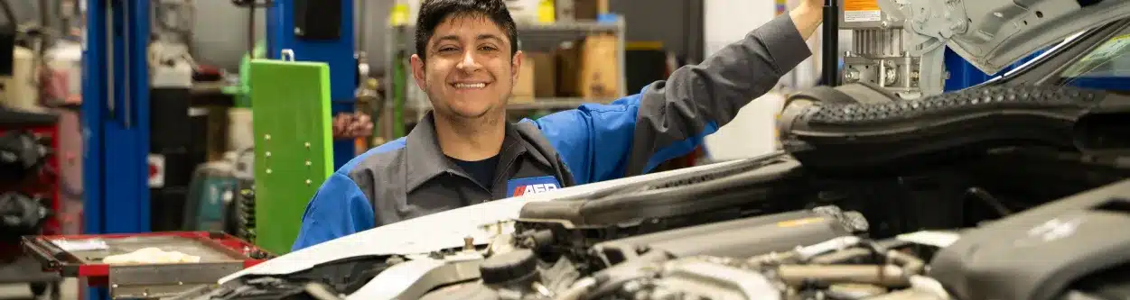 Smiling person in a auto repair shop beside a car with the hood open.